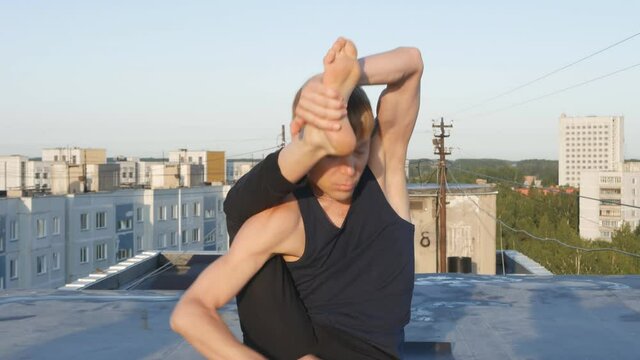 a yogi man put his leg behind his head on his neck and did namaste. Guy yogi at sunset on the roof in summer. Harmony and nature concept.