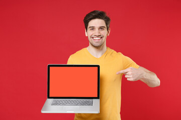 Young smiling unshaved man in basic yellow t-shirt using pointing index finger on laptop pc computer blank screen workspace copy space area mock up isolated on red color background studio portrait