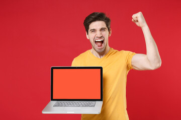 Young happy unshaved happy man in yellow t-shirt using laptop pc computer blank screen workspace area copy space mock up doing winner gesture clenching fist isolated on red background studio portrait