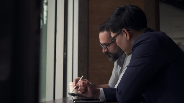4K Two smart business people Caucasian and Asian man sitting by the window together in cafe working and discuss business project ideas with using digital tablet. Businessman teamwork meeting concept.