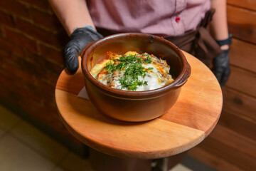 Roasted vegetables and meat under melted cheese in a pot. Baked potatoes dish. Waiter serving dinner at restaurant.