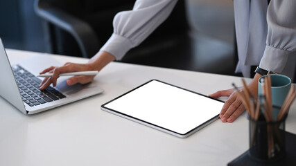 Young female designer using digital tablet and searching information on laptop computer in office.