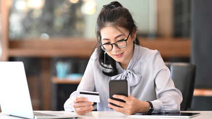 Young woman holding credit card in hand and making payment online or shopping online on website with smart phone in office.