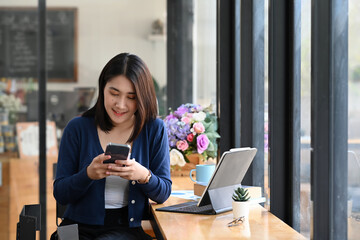 Fototapeta premium Cheerful female chatting online in social networks with friend on smart phone while sitting in office.