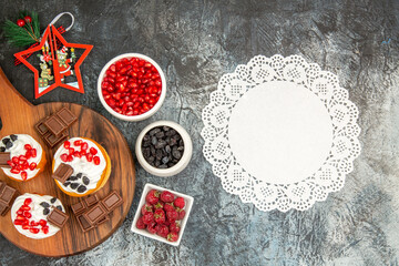 top view berry cakes and chocolates on chopping board xmas toys bowls with pomegranate raisins and raspberries white lace doily on grey-white background