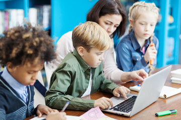 Side view of female teacher explaining group of diverse elementary students  how to study on laptop...