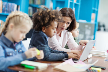 Side view selective focus of female teacher explaining task on laptop screen to mixed race elementary student, his classmates studying sitting at desk in school classroom nearby
