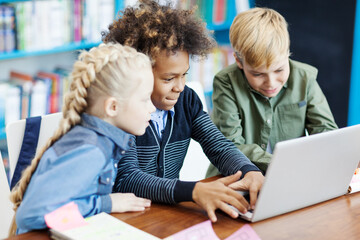 Side view of three curious diverse students with laptop sitting at desk in elementary school classroom. Mixed  race schoolboy playing computer game, his friends supporting him