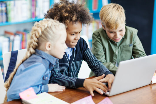 Side view of three curious diverse students with laptop sitting at desk in elementary school classroom. Mixed  race schoolboy playing computer game, his friends supporting him