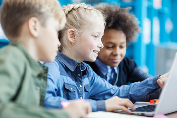 Group of three elementary students sitting at desk in classroom and studying or playing on laptop;...