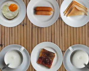Fried egg, sausage, fresh milk, bread spread with jam, bacon on white plate, on bamboo background