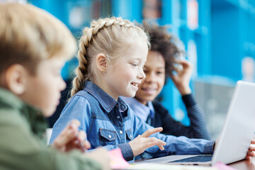 Group of three elementary students sitting at desk in school library and browsing Internet on laptop; focus on smiling blonde girl using touchpad - Powered by Adobe