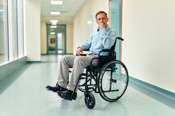 A sad, upset elderly disabled man in a wheelchair sits in the middle of a clinic corridor waiting for his family.