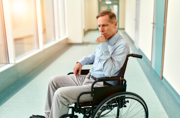 A sad, upset elderly disabled man in a wheelchair sits in the middle of a clinic corridor waiting for his family.