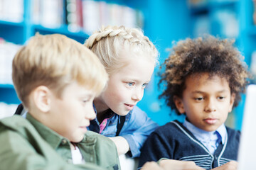 Side view of three curious school friends, two boys and girl, browsing Internet on laptop in school library. Focus on blone girl with braided hair