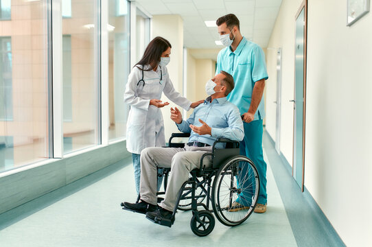 Caucasian Nurse And Handsome Young Doctor Wearing Surgical Masks To Protect Against Covid 19 Pandemic Take Care Of A Mature Male Patient Sitting In A Wheelchair In A Hospital.