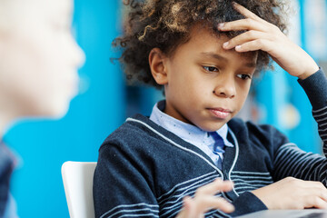Side view of focused mixed race elementary student with curly hair thinking over task with his hand on forehead sitting at desk in school classroom