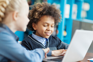 Side view of two smiling kids, boy and girl, using laptop computer together sitting at desk in school library. Focus on curly-haired mixed race male student