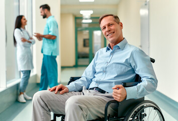 A handsome smiling elderly disabled person in a wheelchair sits in the middle of a clinic corridor with doctors behind him.