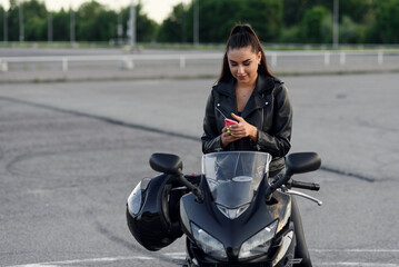 Beautiful female biker uses smartphone while sitting on stylish sport motorcycle at outdoors...