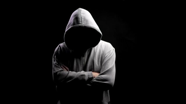 The hooligan provokes. Silhouette of a man with a darkened face in a hood on a black background. A portrait of a murderer, a hacker.