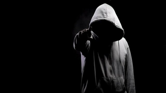 Pointing a finger. Silhouette of a man with a darkened face in a hood on a black background. A portrait of a murderer, a hacker.