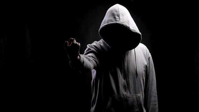 Pointing a finger. Silhouette of a man with a darkened face in a hood on a black background. A portrait of a murderer, a hacker.