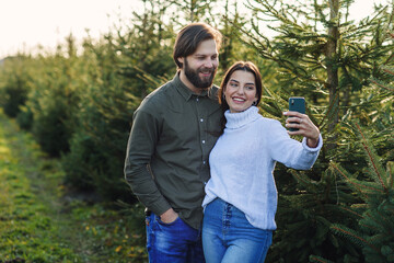 Happy bearded man and his pretty girlfriend making selfie photo at Christmas tree plantation, preparing for the holidays