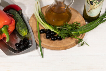 View from above. There is olive oil, green onions, parsley and olives on the cutting board. Nearby is a tray of peppers, cucumbers and olives..