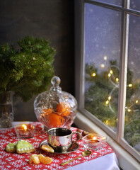 A cup of coffee, tangerines, cookies and fir branches on the table near the snowy window. Lights and candles. Snow outside the window.