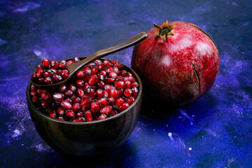 A bowl full of pomegranate seeds with a wooden spoon on it. A whole ripe pomegranate next to the...