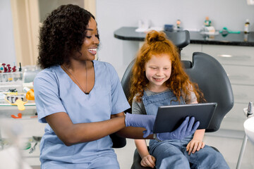 Medicine, pediatric dentistry and oral care concept. Female smiling African dentist showing tablet...