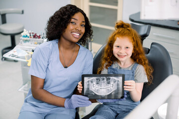Little smiling girl with red curly hair sitting on chair and looking at camera, while holding x-ray scan image of her teeth on digital tablet together with her cheerful black female dentist at clinic