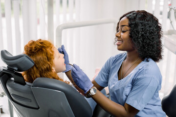 Obraz premium Side view of curly red haired girl lying on dentist chair with open mouth while female African doctor in blue uniform makes dental checkup with tools. Concept of teeth treatment and care.