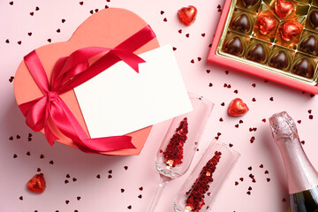 Heart-shaped box with blank paper card, box of chocolates, bottle of champagne and glasses with confetti on pink background. Valentine's day dinner, love, romance concept. Flat lay, top view.