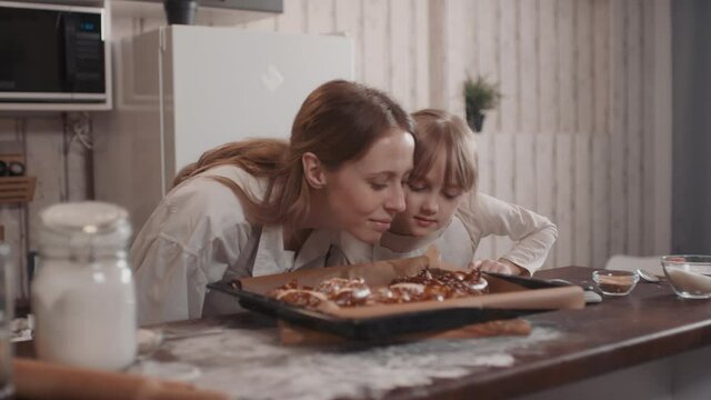 Waist-up shot of happy young caucasian woman bringing hot pretzels on baking sheet and little girl clapping smelling delicious baked food