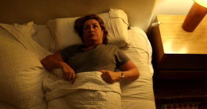 Sleepless senior woman turning light on and rubbing face with hands unable to sleep