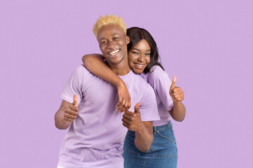 Funky black guy and his girlfriend hugging and showing thumbs up, recommending something over lilac studio background