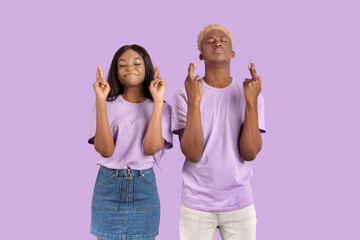 Young black couple crossing fingers and making wish with closed eyes, begging for dream to come true on lilac background