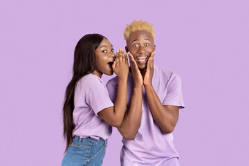 Excited black woman whispering something into her boyfriend's ear over lilac studio background