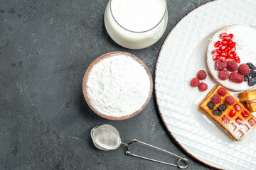 top view tart and waffles with berries on white oval plate a bowl of powdered sugar a sieve and a glass of milk on dark background with copy space
