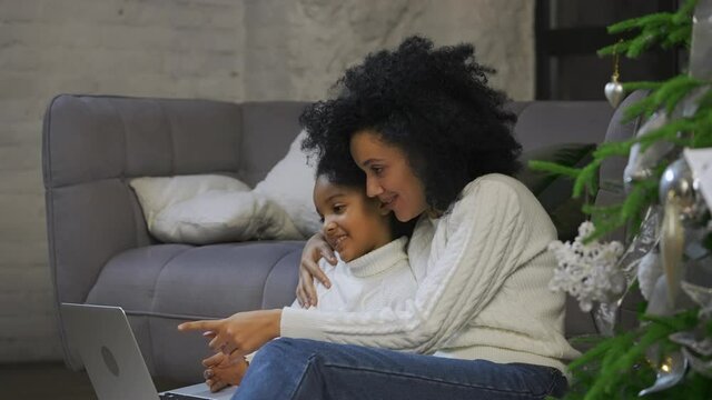 Portrait of African American mom and her little daughter communicate by video call using a laptop. Woman and girl sitting near sofa and decorated Christmas tree. Close up. Slow motion.