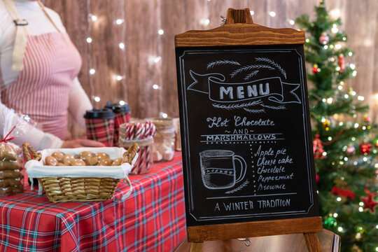 Christmas pastry shop . Woman seller, waitress selling gingerbread, marshmallows, cookies, sweets and hot chocolate in a small cozy cafe. Homemade bakery menu in the foreground