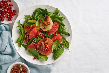 Fruits citrus salad with nuts, green lettuce leaves. Copy space. Balanced food. Spinach with orange, grapefruit, pecans and pomegranate seeds in bowl on table with white tablecloth.