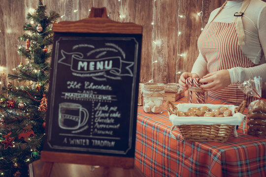 Christmas pastry shop . Woman seller, waitress selling gingerbread, marshmallows, cookies, sweets and hot chocolate in a small cozy cafe. Homemade bakery menu in the foreground