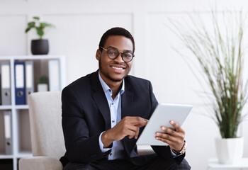 Smiling black manager in suit holding digital tablet, office interior
