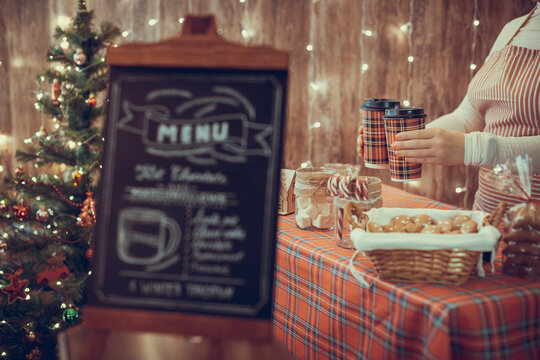Christmas pastry shop . Woman seller, waitress selling gingerbread, marshmallows, cookies, sweets and hot chocolate in a small cozy cafe. Homemade bakery menu in the foreground