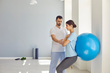 Physiotherapist helping young woman do wall squats with fit ball in modern clinic
