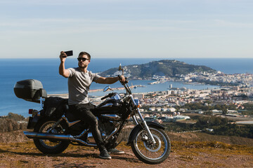 Fototapeta premium Biker sitting on the motorcycle taking selfie with the landscape with the mobile
