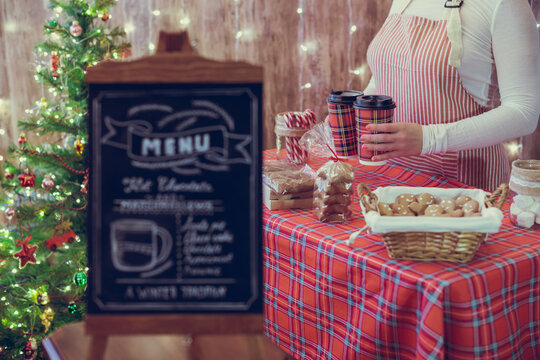 Christmas pastry shop . Woman seller, waitress selling gingerbread, marshmallows, cookies, sweets and hot chocolate in a small cozy cafe. Homemade bakery menu in the foreground